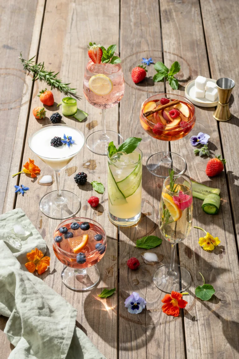 An overhead shot of several white wine cocktails in stemmed glasses on a wooden outdoor table, garnished with fruit and herbs, soft afternoon light, condensation on the glasses